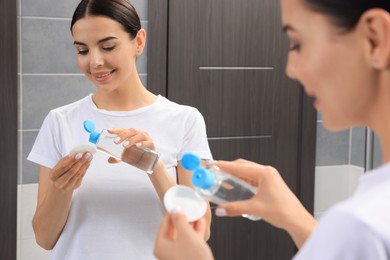Photo of Young woman pouring micellar water onto cotton pad near mirror in bathroom