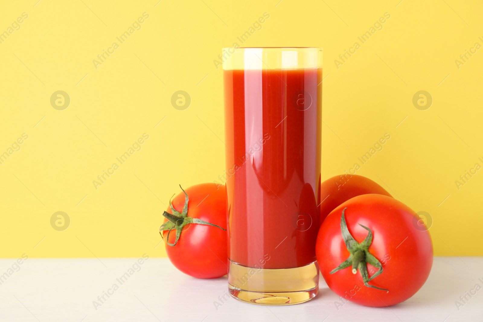 Photo of Glass of delicious juice and fresh tomatoes on white table against yellow background, space for text