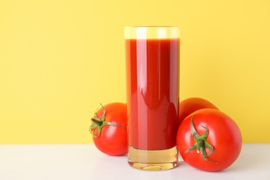 Glass of delicious juice and fresh tomatoes on white table against yellow background, space for text