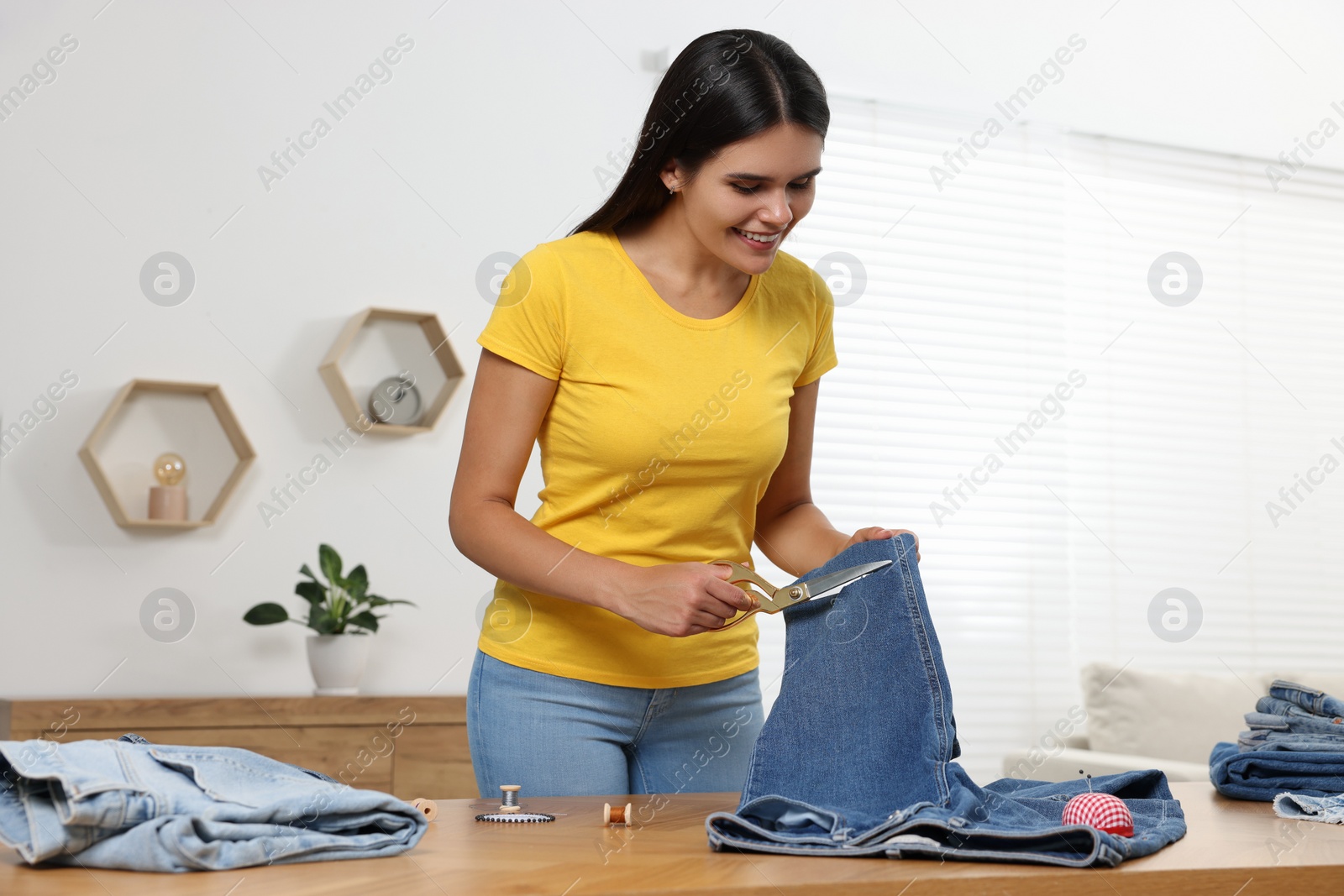 Photo of Young woman cutting jeans with scissors at wooden table indoors