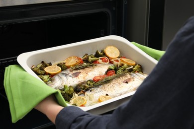 Photo of Woman taking baking dish with delicious fish and vegetables from oven, closeup