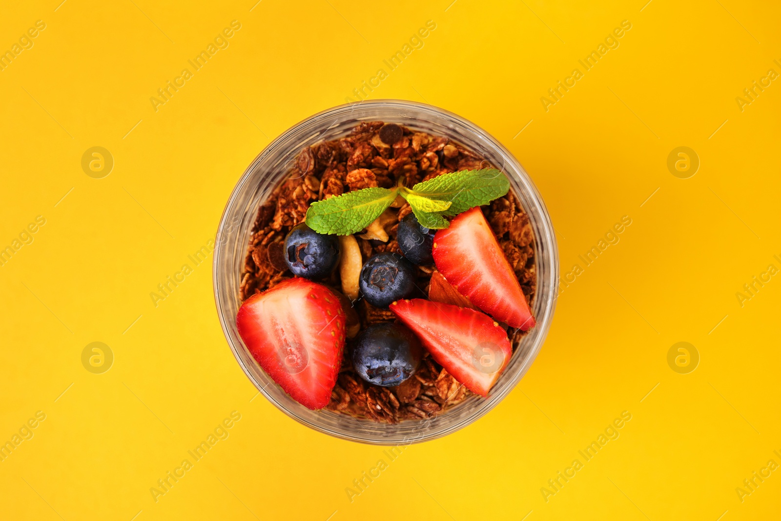 Photo of Tasty granola with berries and mint in glass on yellow background, top view