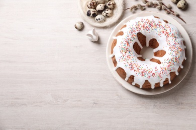 Photo of Glazed Easter cake with sprinkles, decorative bunny, quail eggs and willow branches on white wooden table, flat lay. Space for text