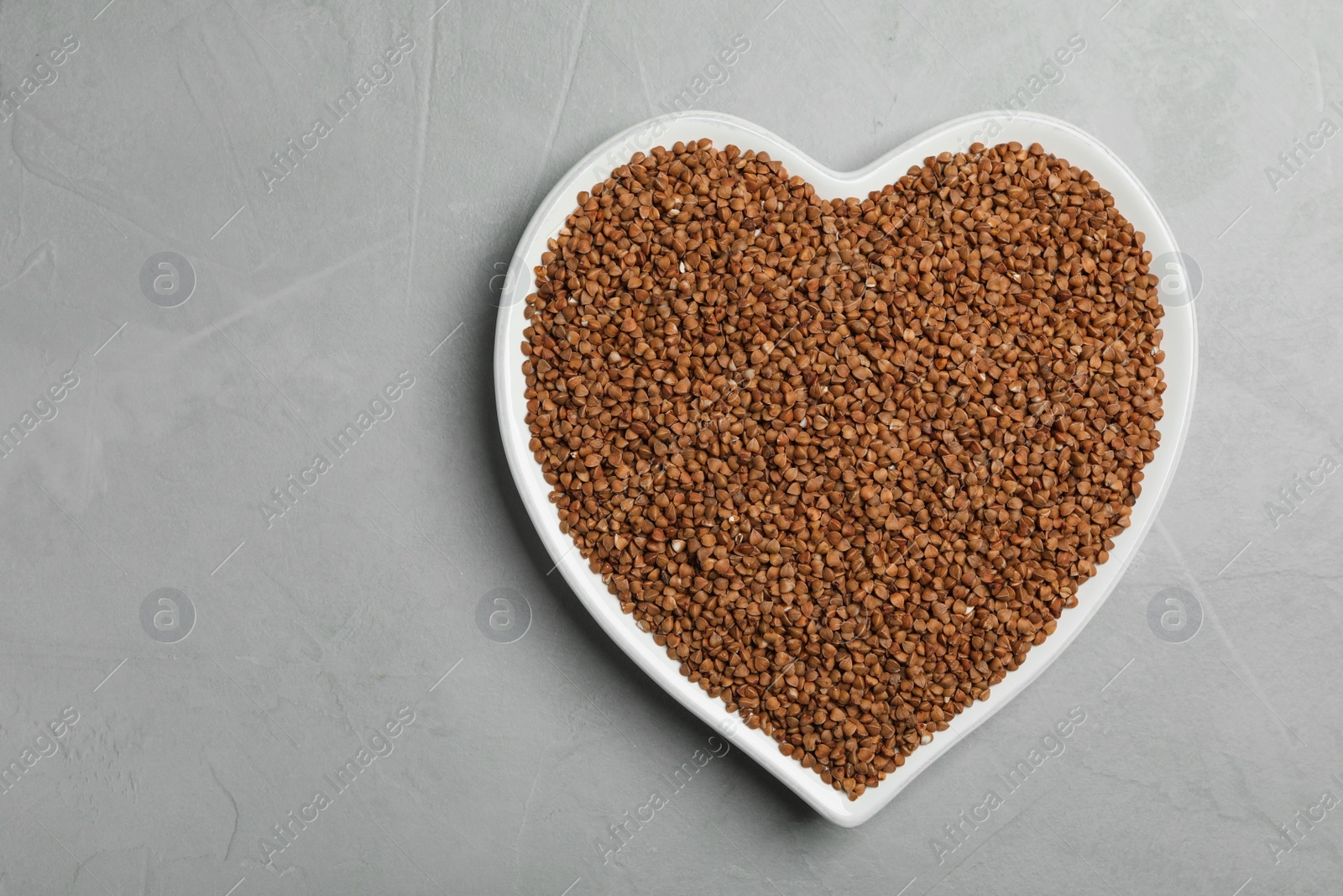 Photo of Heart shaped plate with raw buckwheat on grey background, top view. Space for text