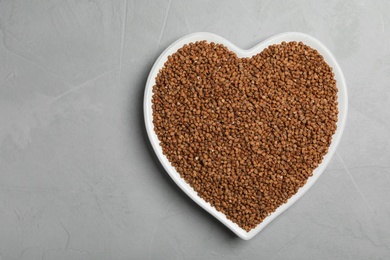 Heart shaped plate with raw buckwheat on grey background, top view. Space for text