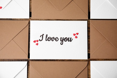 Sheet of paper with phrase I Love You, envelopes and heart shaped sprinkles on wooden table, flat lay