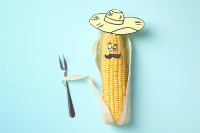 Photo of Mexican man made of corncob, paper hat and fork on light blue background, top view