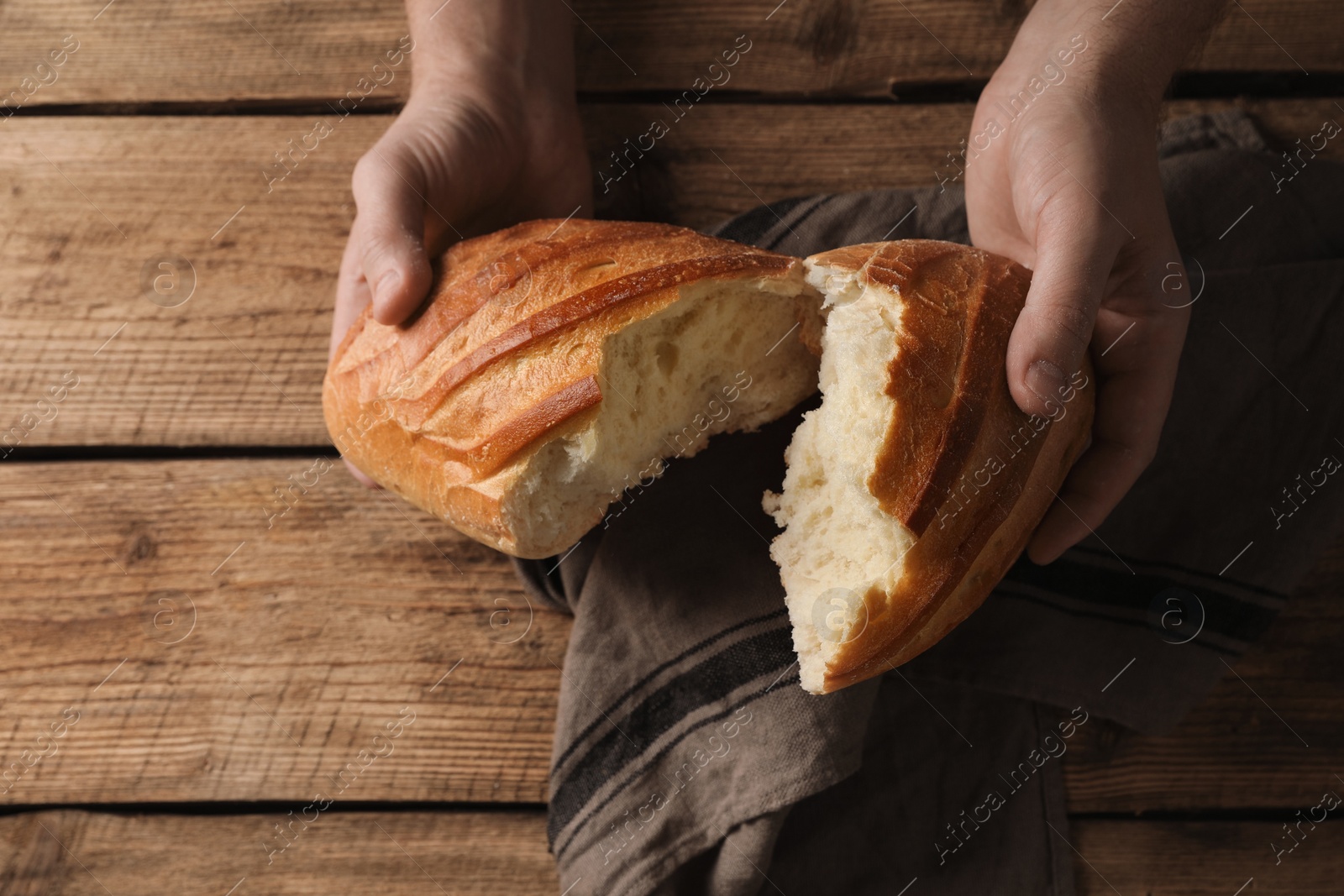 Photo of Man breaking loaf of fresh bread at wooden table, top view