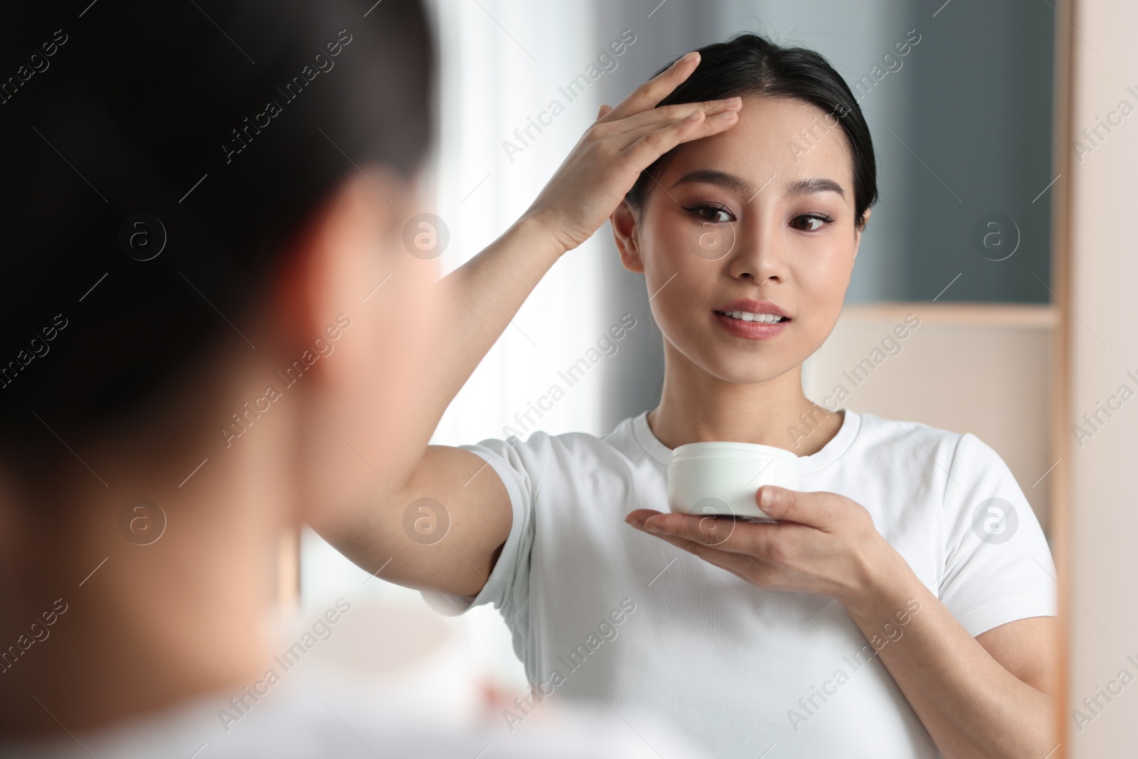 Photo of Happy woman applying face cream near mirror at home