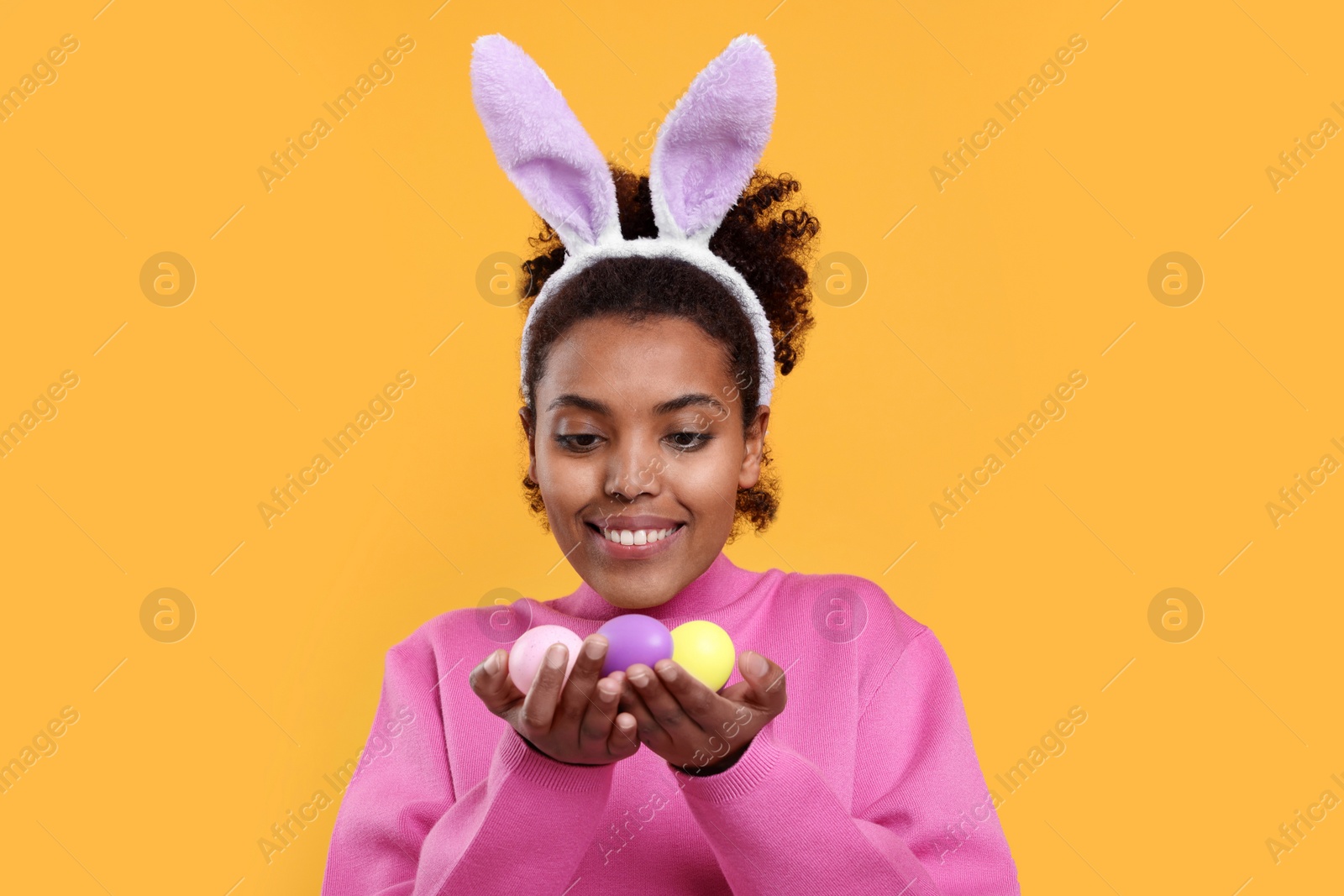 Photo of Happy African American woman in bunny ears headband holding Easter eggs on orange background