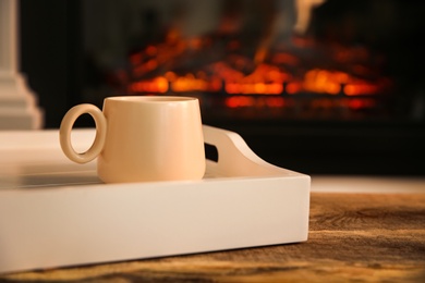 Photo of Cup with hot drink in tray on wooden table against fireplace, space for text