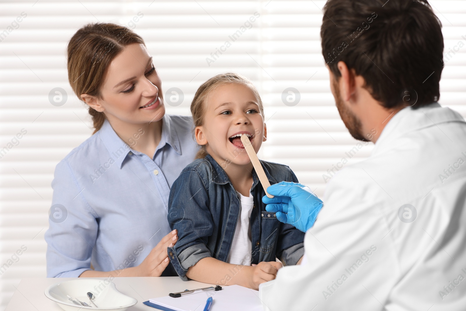 Photo of Doctor examining girl`s oral cavity with tongue depressor near her mother indoors