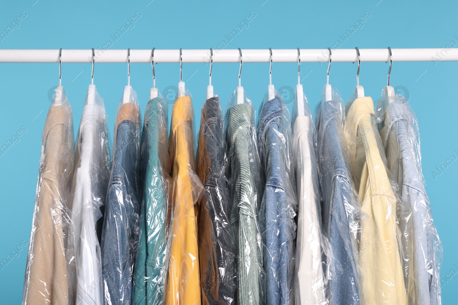 Photo of Dry-cleaning service. Many different clothes in plastic bags hanging on rack against light blue background