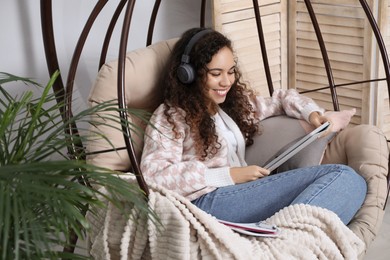 Photo of African American woman with headphones and tablet studying in egg chair at home. Distance learning