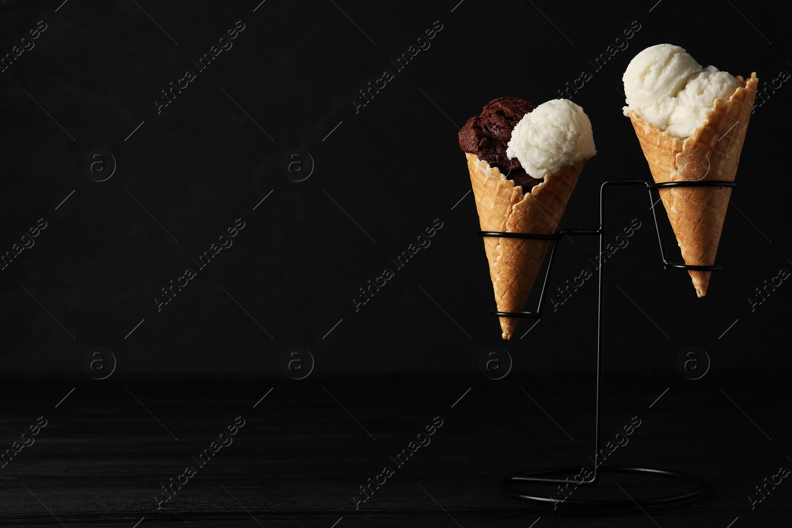 Photo of Ice cream scoops in wafer cones on black wooden table against dark background, space for text