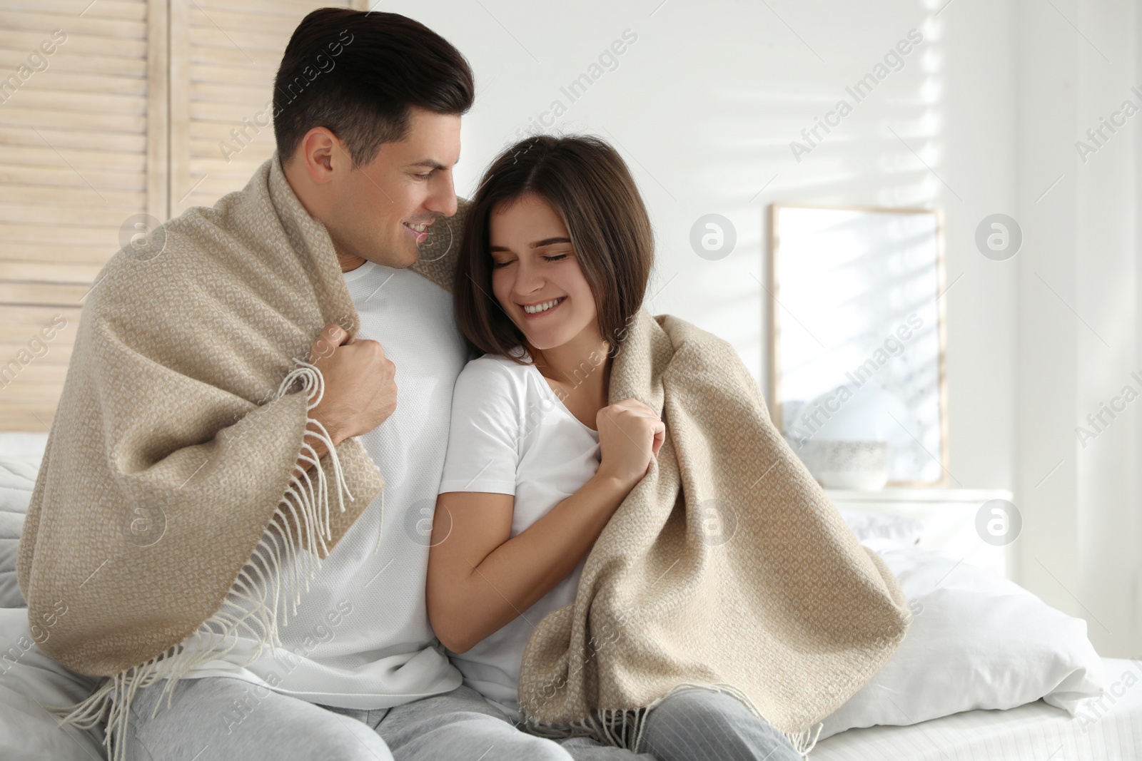 Photo of Happy couple covered with warm beige plaid sitting on bed indoors