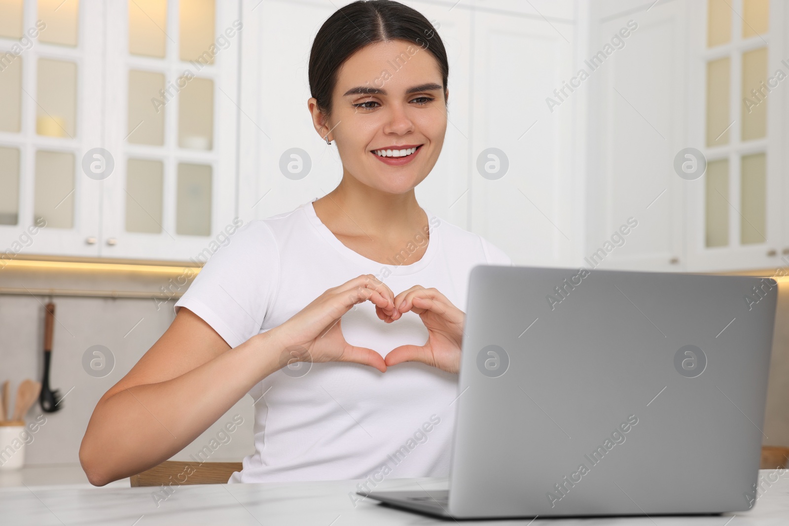 Photo of Happy young woman having video chat via laptop and making heart at table in kitchen