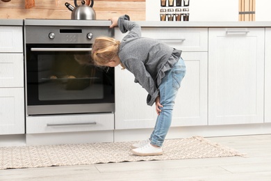Photo of Little girl waiting for preparation of cookies in oven at home