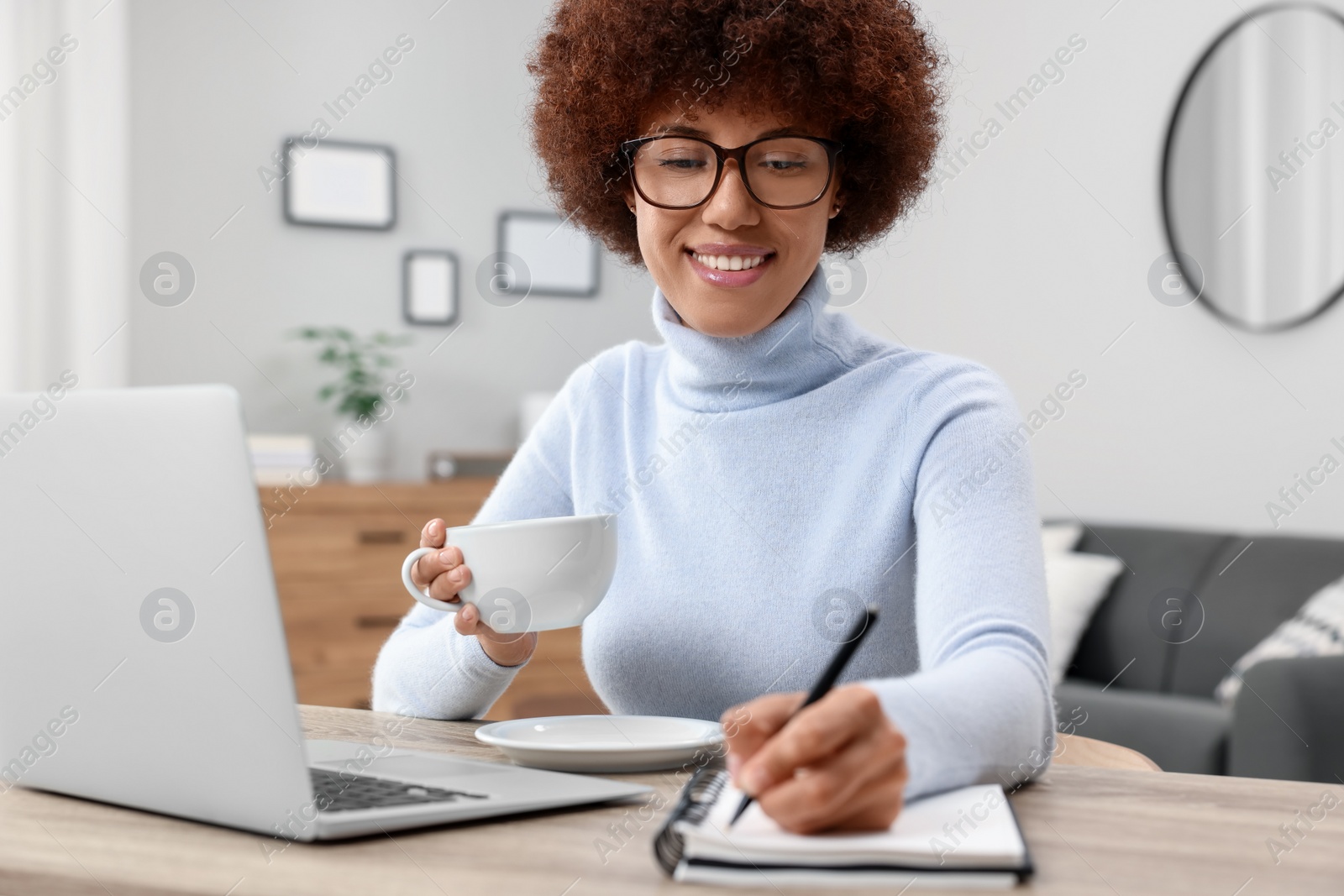 Photo of Beautiful young woman writing in notebook near laptop at wooden desk in room