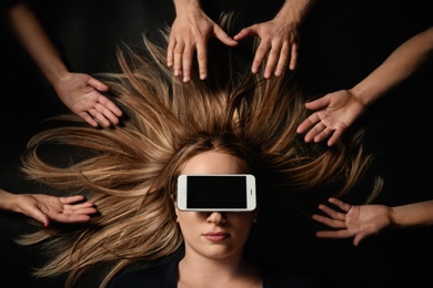 Young woman with smartphone covering her eyes surrouded by people's hands on black background, top view. Space for text
