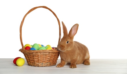 Photo of Cute bunny and basket with Easter eggs on table against white background