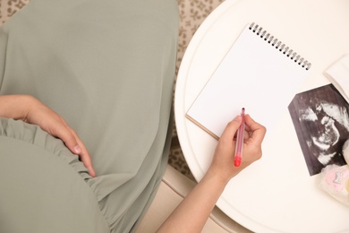 Photo of Pregnant woman with sonogram writing baby names list at small white table, top view