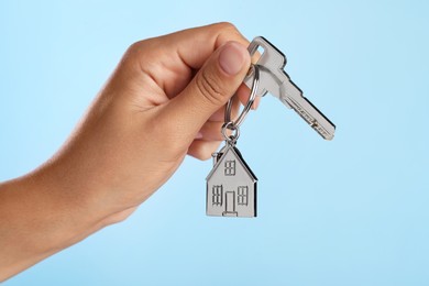 Photo of Woman holding key with metallic keychain in shape of house on light blue background, closeup