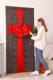 Photo of Woman holding Christmas tree and decorating wooden door with red bow indoors