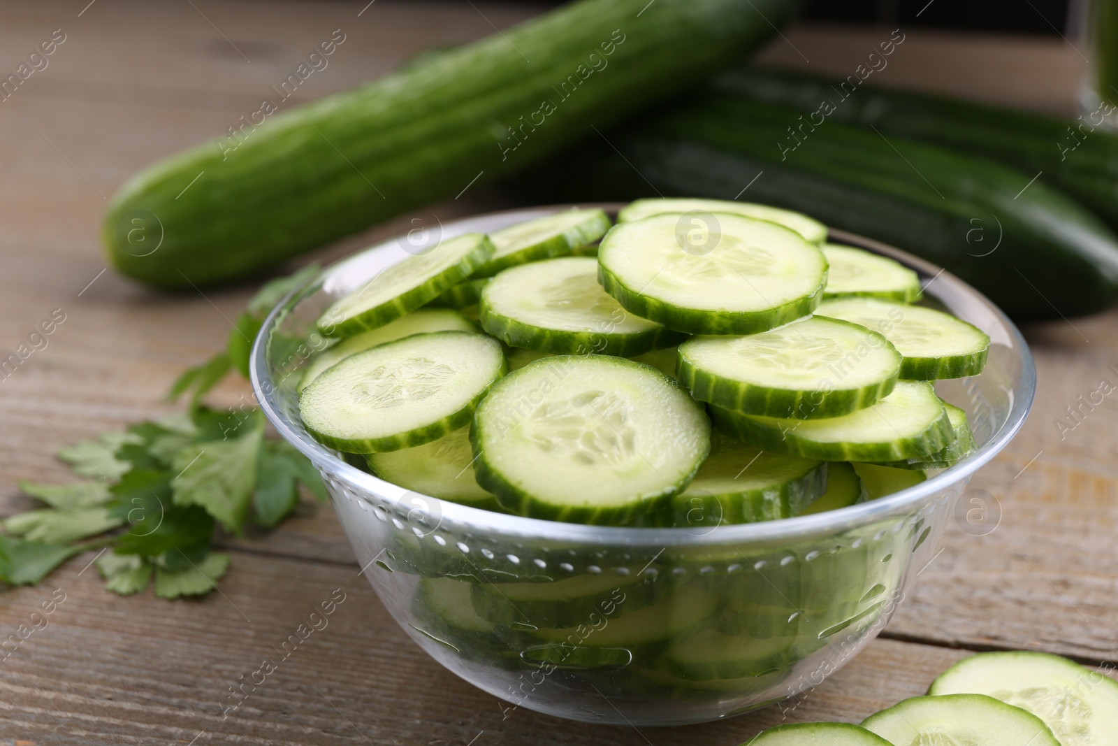 Photo of Cut cucumber in glass bowl, fresh vegetables and parsley on wooden table, closeup