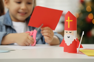 Photo of Cute little girl cutting paper at table, focus on Saint Nicholas toy