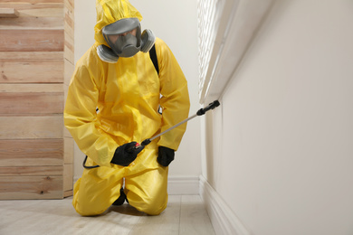 Photo of Pest control worker in protective suit spraying insecticide on window sill at home