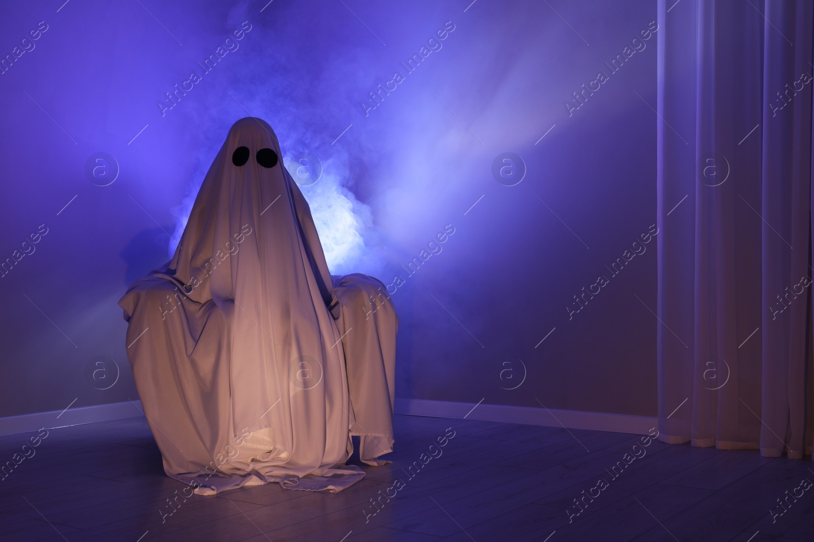 Photo of Creepy ghost. Woman covered with sheet sitting in armchair in blue light, space for text