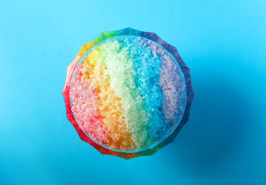 Rainbow shaving ice in glass dessert bowl on light blue background, top view