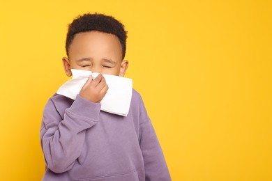 Photo of African-American boy blowing nose in tissue on yellow background, space for text. Cold symptoms