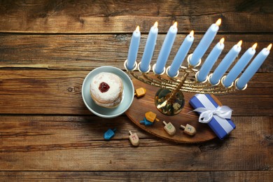 Photo of Hanukkah celebration. Menorah with burning candles, dreidels, donuts and gift box on wooden table, above view. Space for text