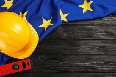 Photo of Yellow hard hat, building level and flag of European Union on black wooden table, flat lay. Space for text