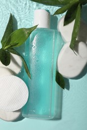 Wet bottle of micellar water, cotton pads and green twigs on light blue background, flat lay