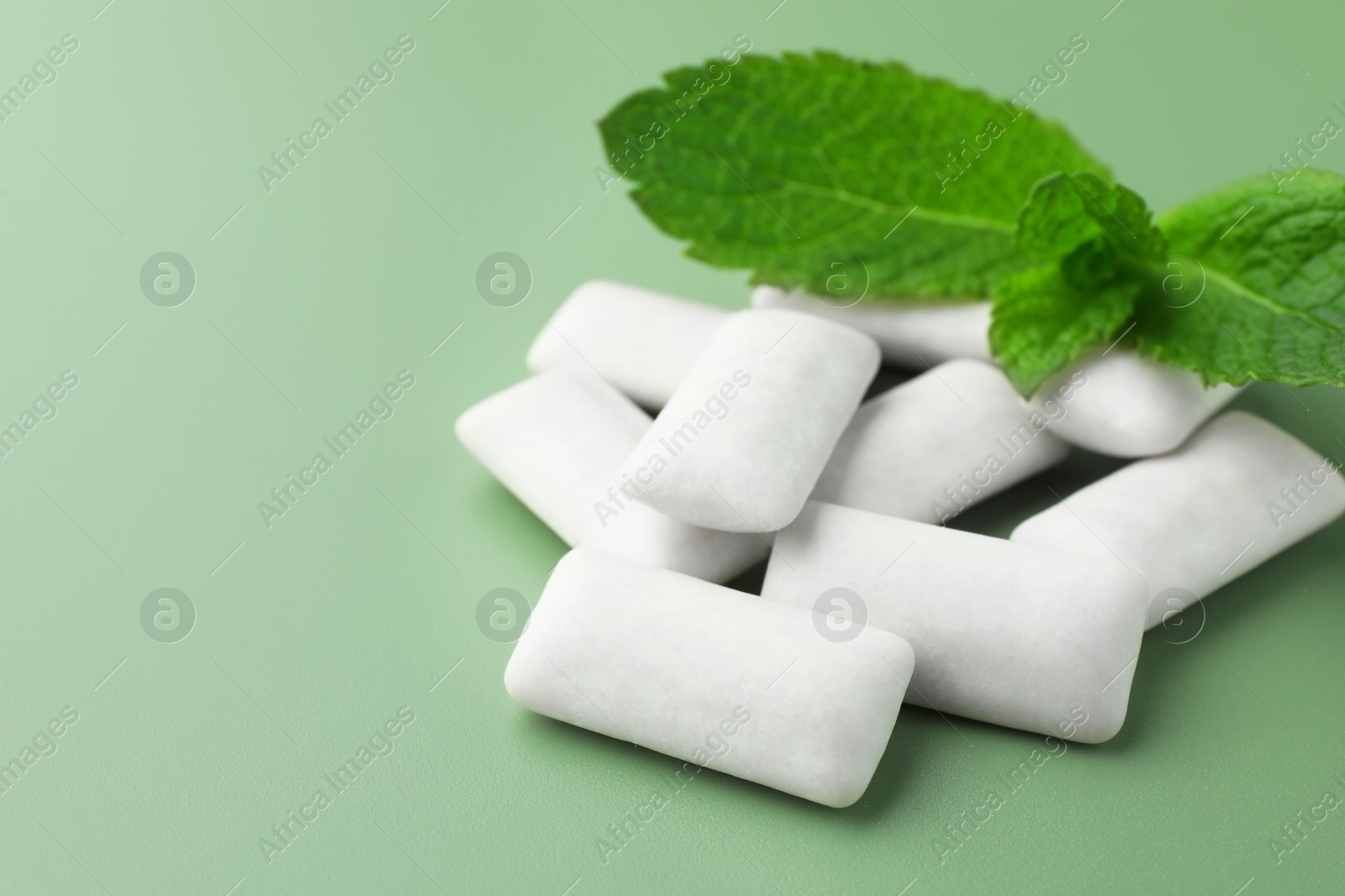 Photo of Tasty white chewing gums and mint leaves on light green background, closeup. Space for text