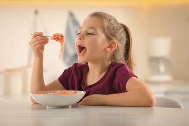 Photo of Cute little girl eating tasty pasta at table in kitchen