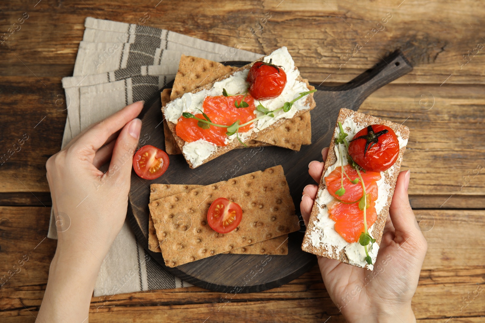 Photo of Woman eating fresh rye crispbreads with salmon, cream cheese and tomatoes at wooden table, top view