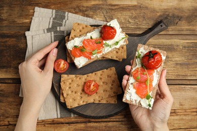 Photo of Woman eating fresh rye crispbreads with salmon, cream cheese and tomatoes at wooden table, top view