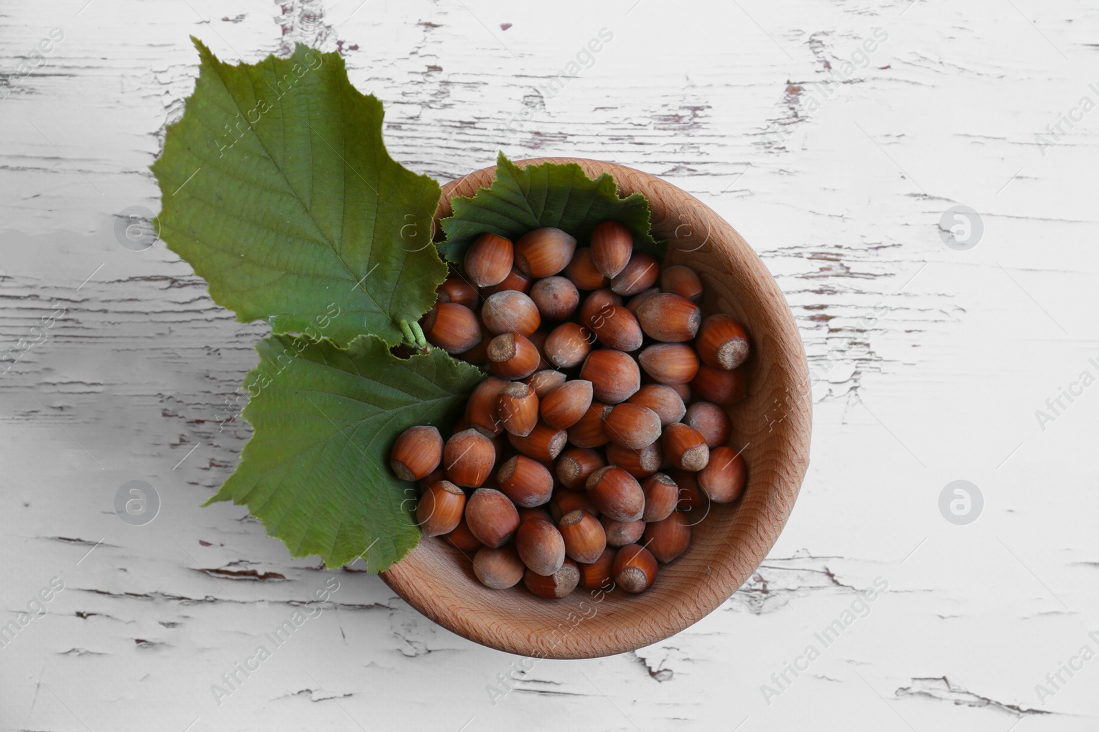 Photo of Bowl with hazelnuts and leaves on white wooden table, top view