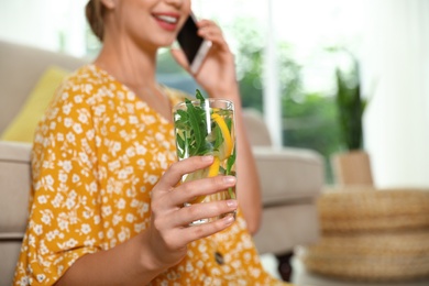 Young woman with glass of lemonade talking on phone at home, closeup. Refreshing drink