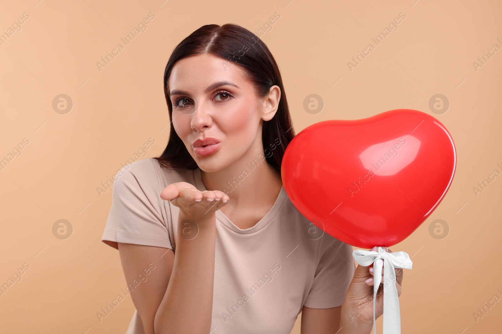 Photo of Young woman holding red heart shaped balloon and blowing kiss on beige background
