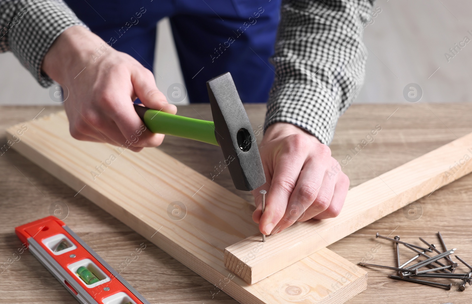 Photo of Professional repairman hammering nail into board at wooden table indoors, closeup