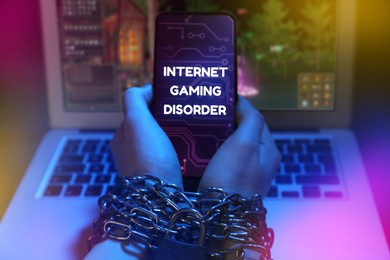 Image of Internet gaming disorder. Woman with chained hands holding smartphone near laptop on dark background, closeup