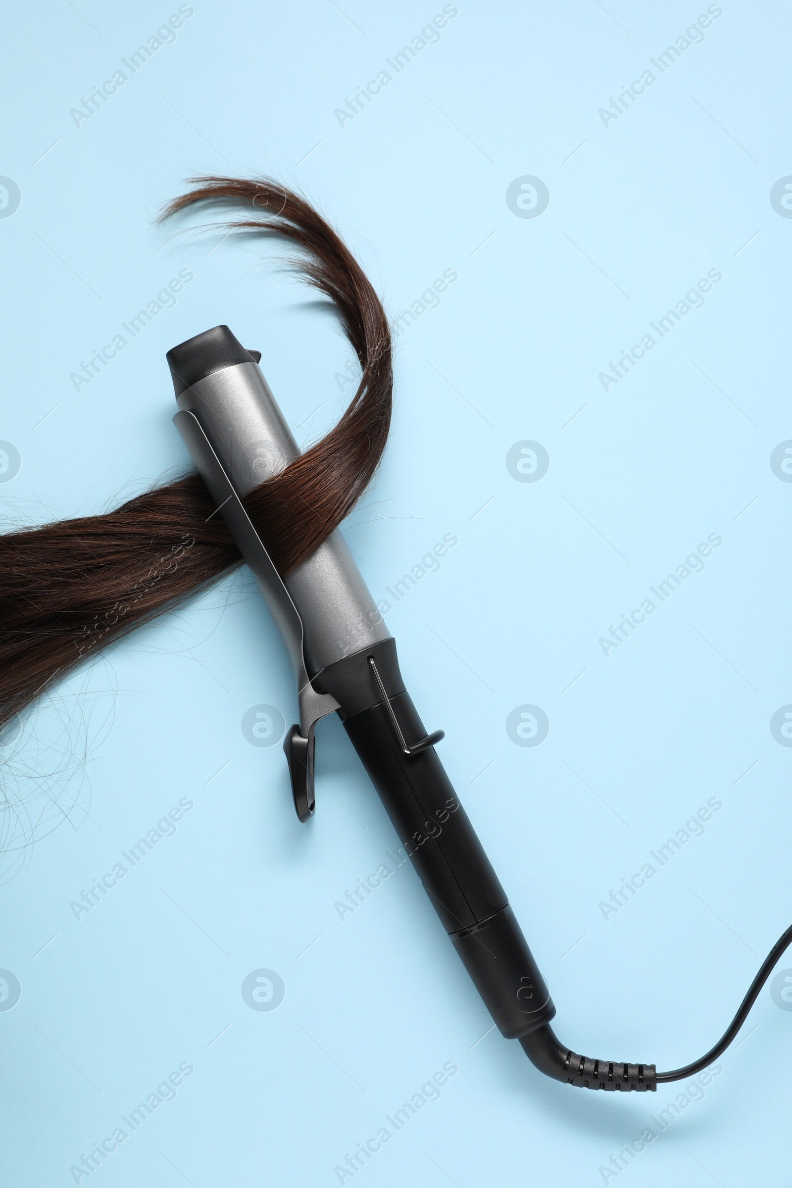 Photo of Curling iron with brown hair lock on light blue background, top view