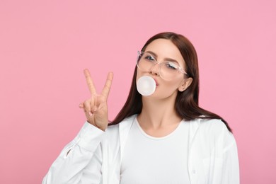 Beautiful woman in glasses blowing bubble gum and showing V-sign on pink background