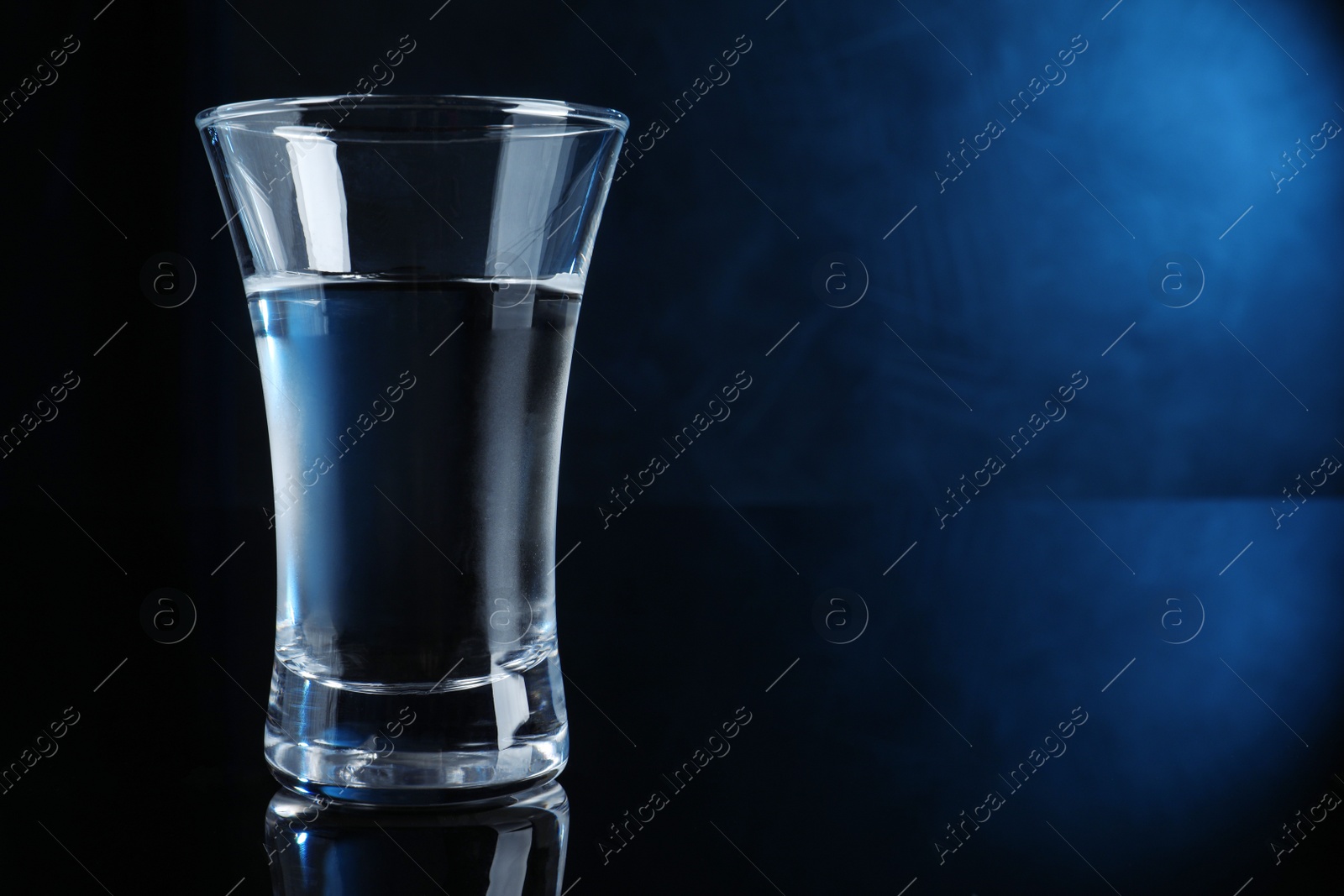 Photo of Shot glass of vodka on dark background with blue light. Space for text