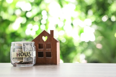 Glass jar with money and house model on table against blurred background. Space for text